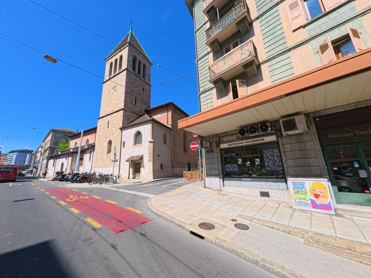 Eine Kirche mit Uhrturm erhebt sich in Genève (1201, Kanton Genf) entlang einer Strasse mit Fahrradspur und angrenzenden Wohnhäusern.