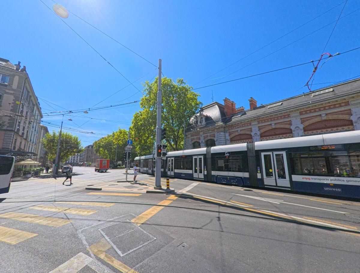 Eine moderne Strassenbahn fährt in Genève (1201, Kanton Genf) an einem historischen Gebäude vorbei, während Passanten die sonnige Kreuzung überqueren.