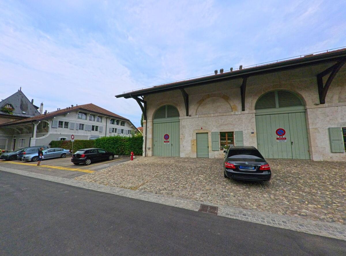 Ein langgestrecktes Gebäude mit grossen Torbögen und grünen Fensterläden steht in Genthod (1294, Kanton Genf) neben einem kleinen Parkplatz.