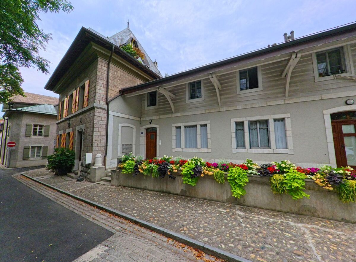 Ein historisches Gebäude mit bunten Fensterläden und Blumenschmuck prägt eine Kopfsteinpflastergasse in Genthod (1294, Kanton Genf).