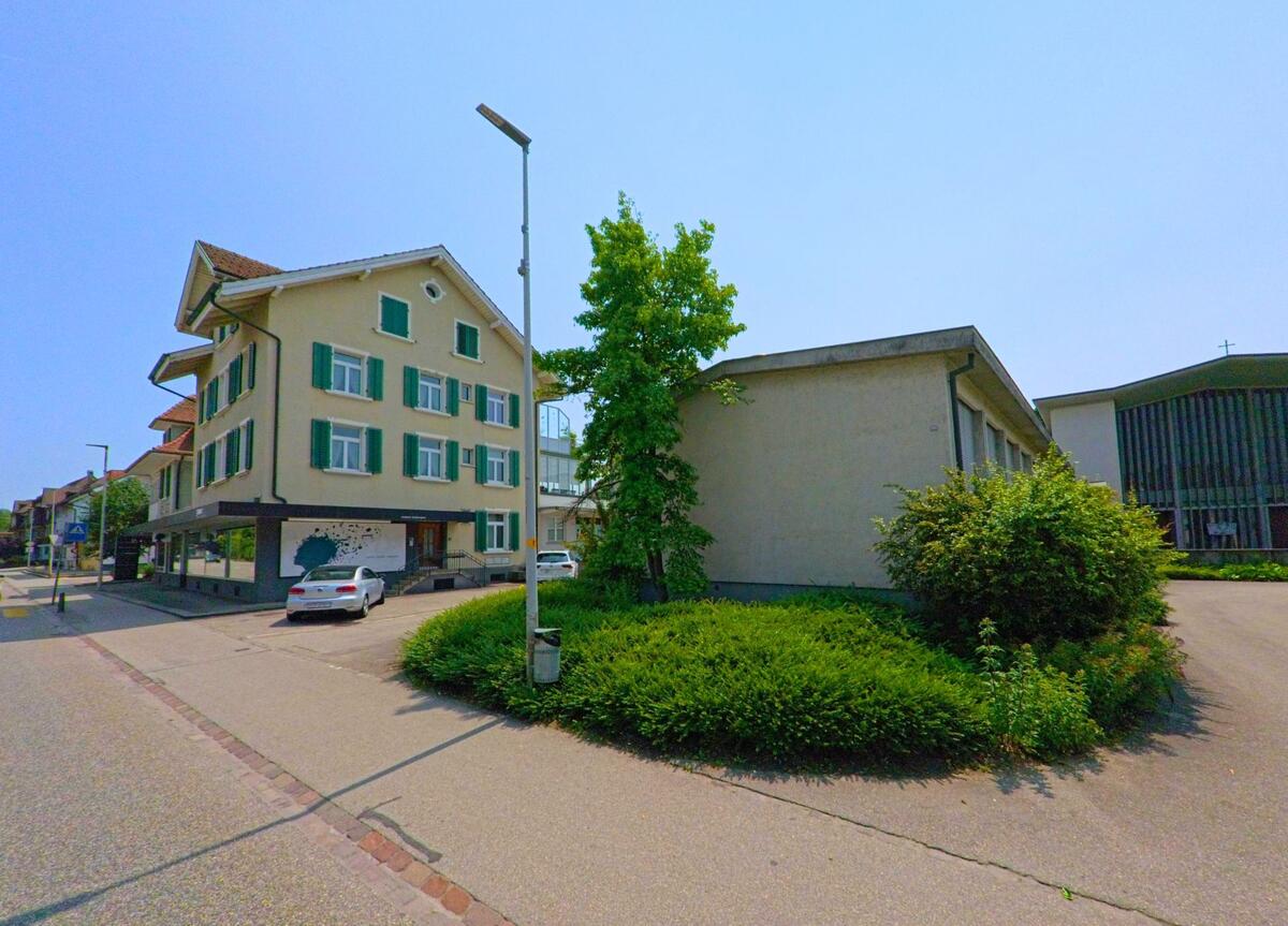  In Gerlafingen (4563, Kanton Solothurn) steht ein Wohn- und Geschäftshaus mit grünen Fensterläden gegenüber einer modernen Kirche mit geschwungener Fassade.