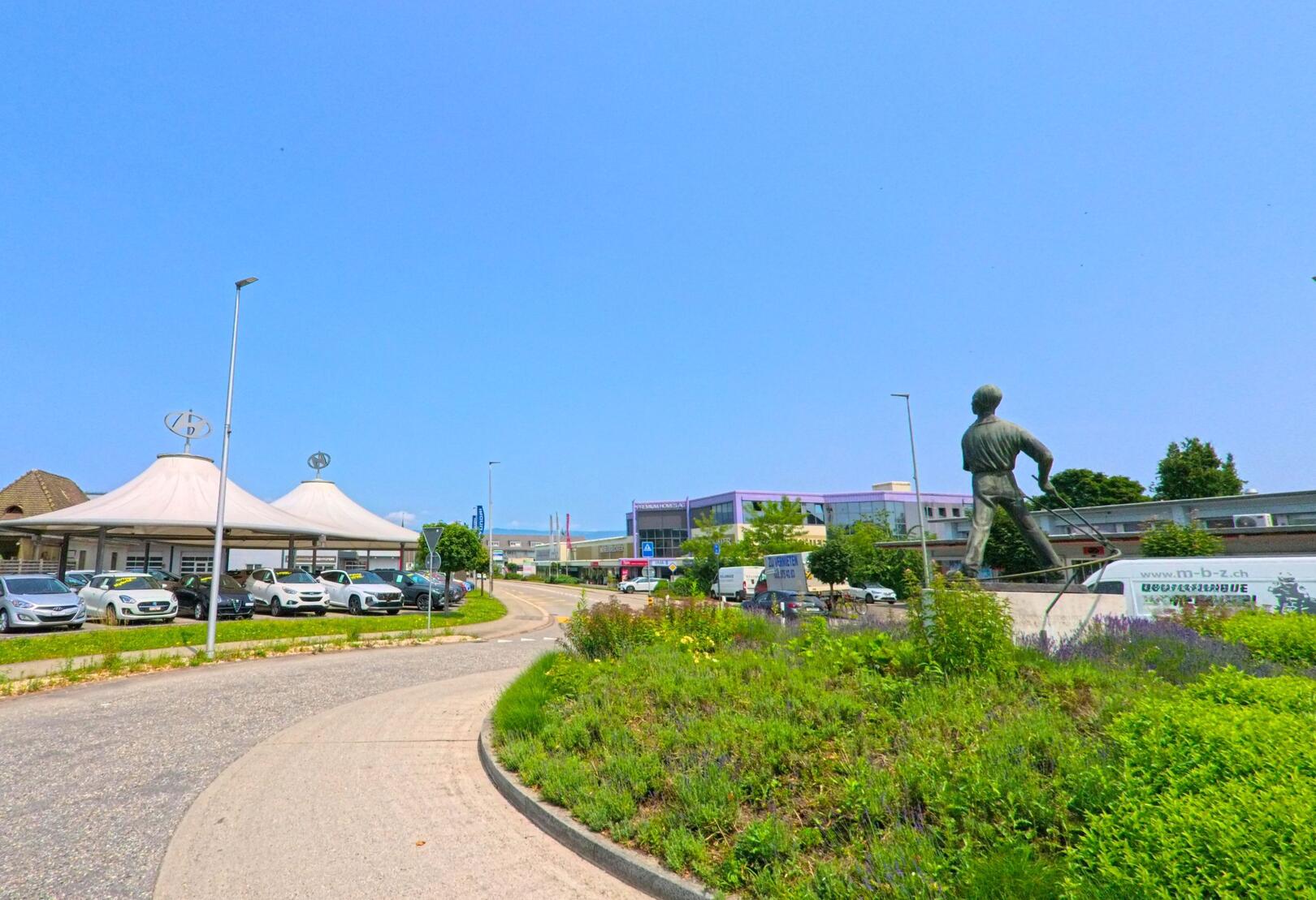 Am Rand eines Kreisverkehrs in Gerlafingen (4563, Kanton Solothurn) steht eine markante Bronzestatue, die in Richtung eines modernen Gewerbegebiets mit Autohaus und Industriehallen blickt.