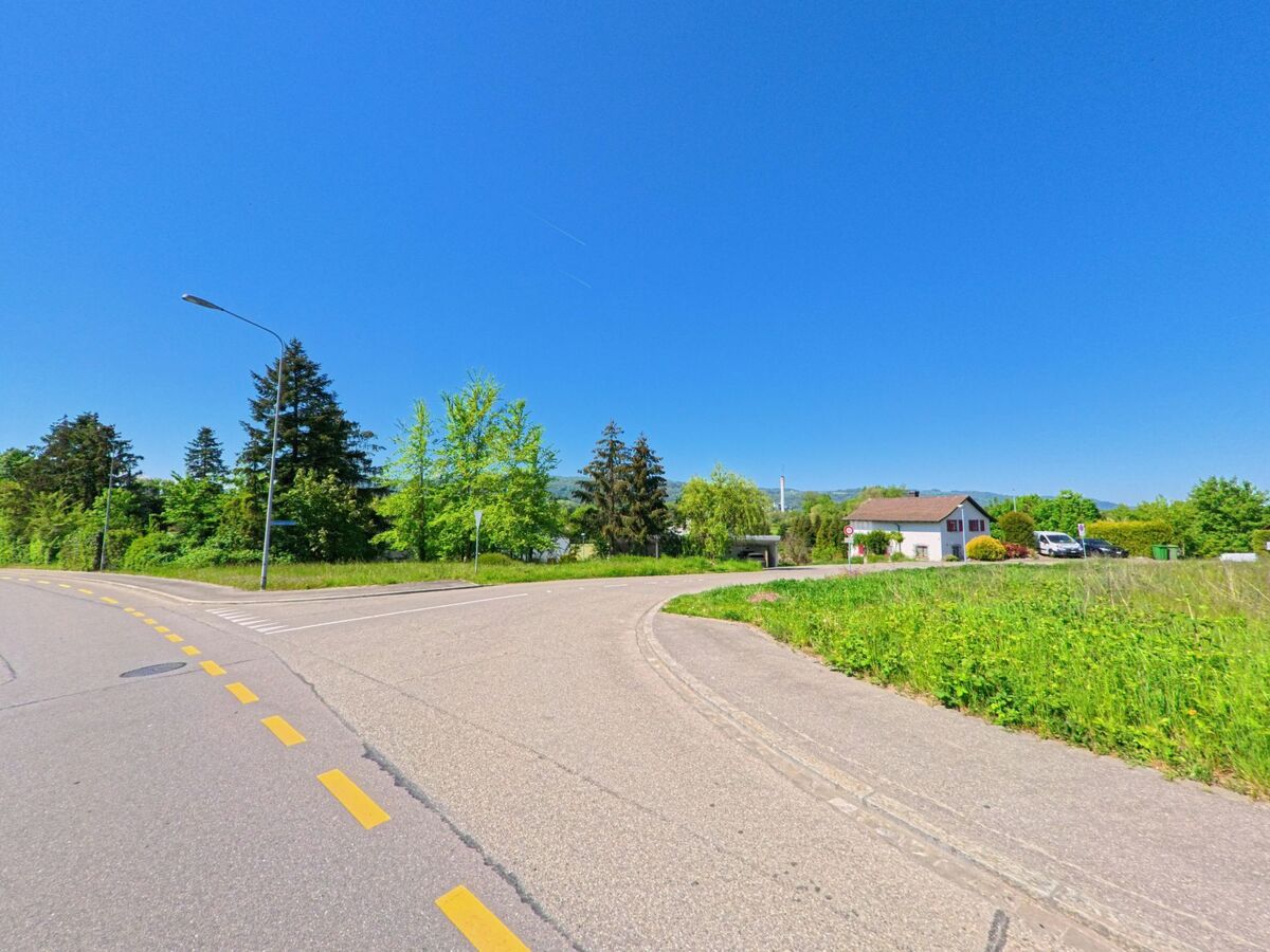 In Geroldswil (8954, Kanton Zürich) ist eine kurvige Strasse zu sehen, die an einem grünen Feld und einem Einfamilienhaus vorbei führt, während der klare Himmel und die Bäume die Szene ergänzen.