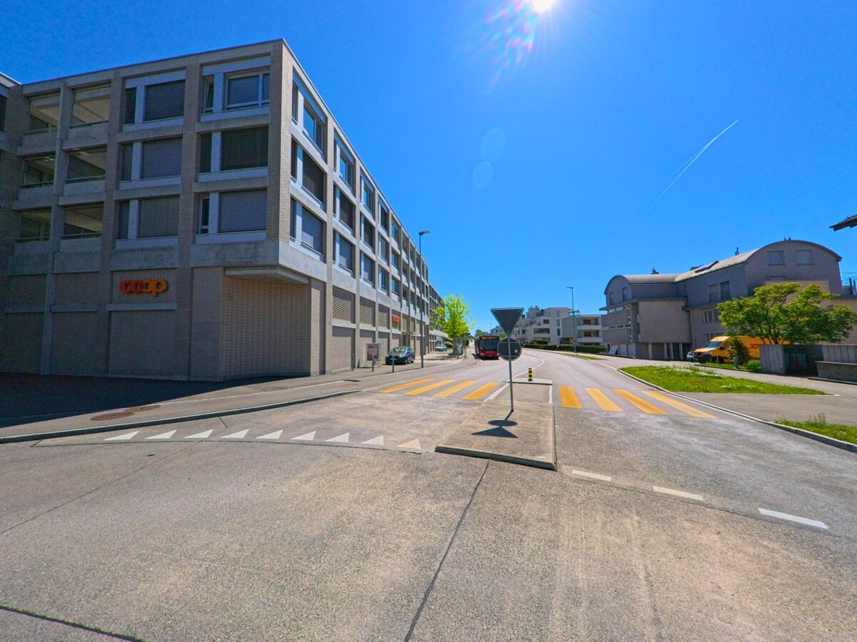 An einem sonnigen Tag führt eine Strasse in Geroldswil im Kanton Zürich mit der PLZ 8954 an einem Coop-Gebäude vorbei, flankiert von modernen Wohnhäusern und einem Fussgängerstreifen.