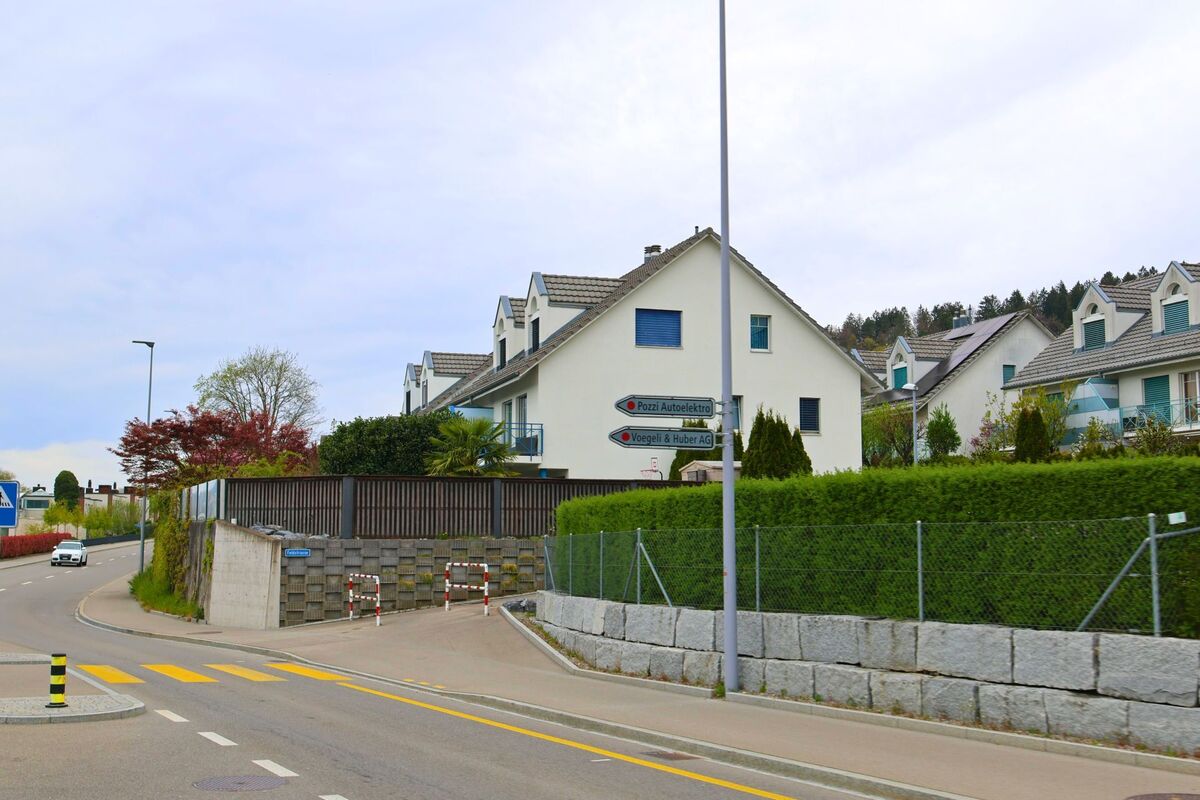 In Geroldswil im Kanton Zürich mit der PLZ 8954 führen Wegweiser an einer Strassenkreuzung zu lokalen Unternehmen, während Reihenhäuser das Bild am Hang prägen.