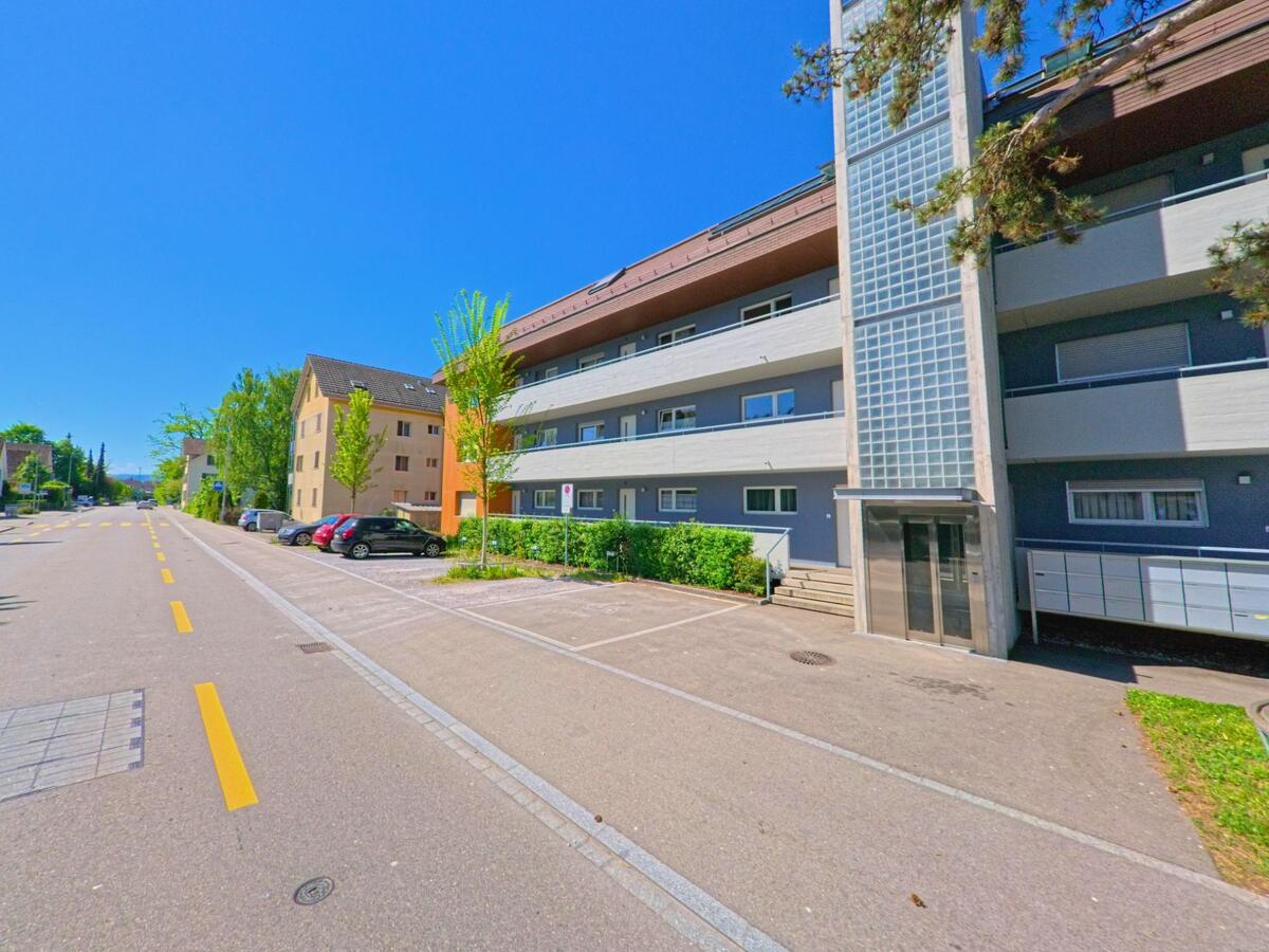 In Geroldswil im Kanton Zürich mit der PLZ 8954 säumen mehrstöckige Wohnhäuser eine ruhige Strasse mit markierten Parkplätzen und jungen Bäumen bei sonnigem Wetter.