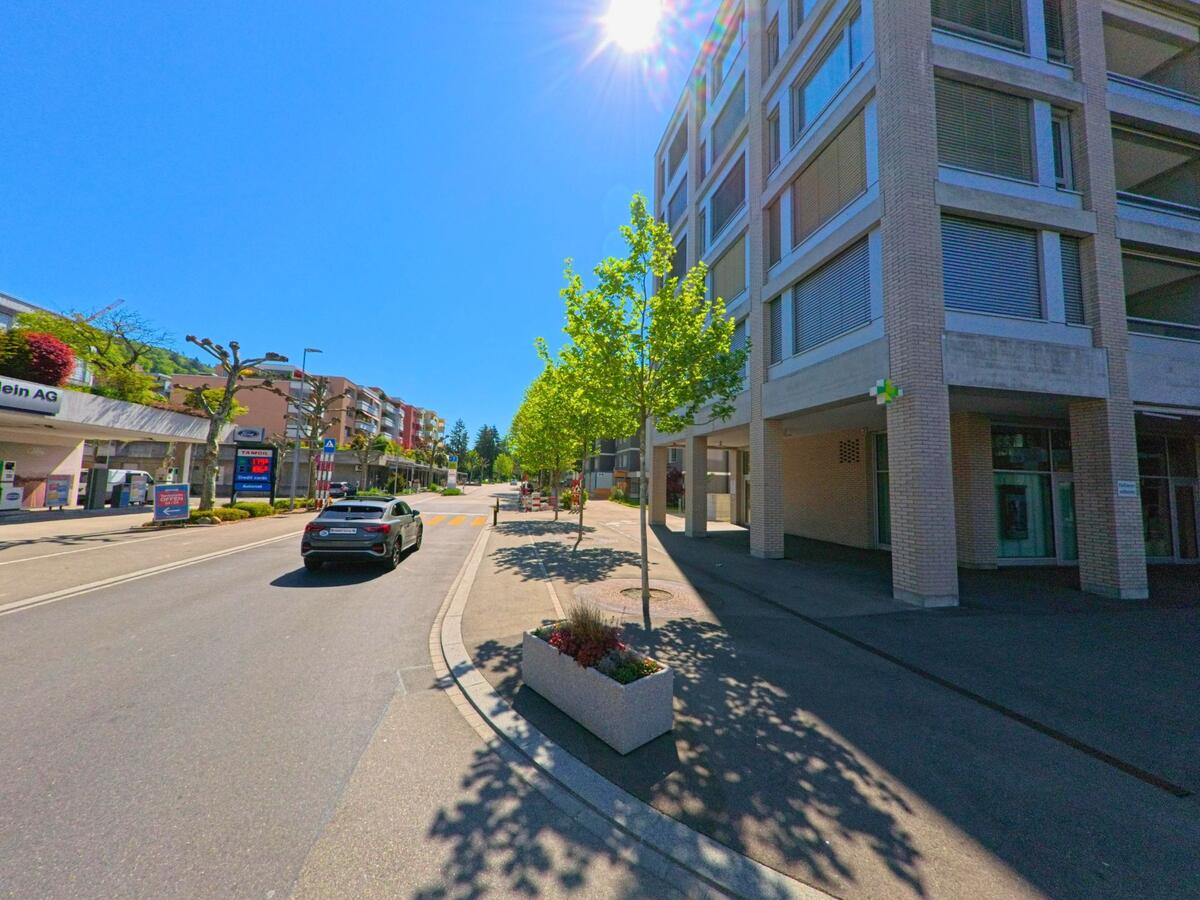 In der Gemeinde Geroldswil im Kanton Zürich mit der PLZ 8954 verläuft eine zentrale Strasse mit modernen Gebäuden, einer Apotheke und einer nahegelegenen Tankstelle bei strahlendem Sonnenschein.
