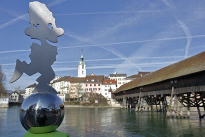 Vor der Holzbruecke in Olten (4600, Kanton Solothurn) steht eine metallene Figur auf einer Kugel, waehrend im Hintergrund die Altstadt und die Kirche zu sehen sind.