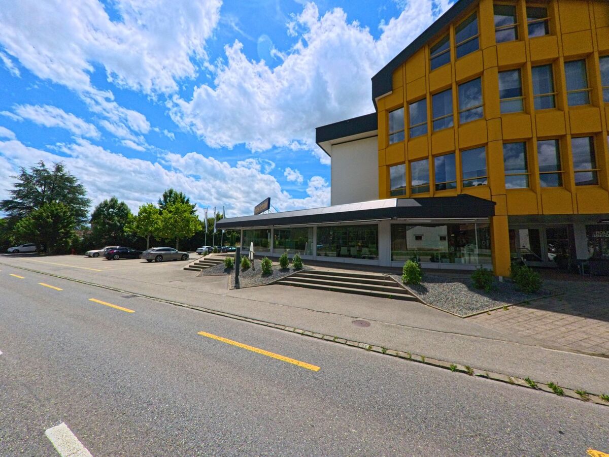  In Geuensee (6232, Kanton Luzern) steht ein markantes Gebaeude mit gelber Fassade und grossen Fenstern an einer Strasse mit vorgelagertem Parkplatz und Treppenaufgang.