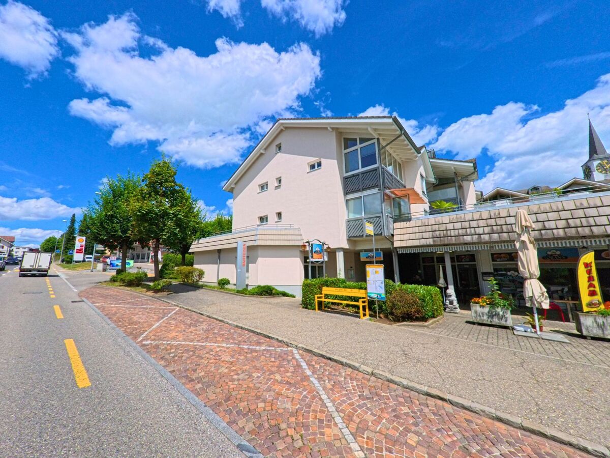 Im Zentrum von Geuensee (6232, Kanton Luzern) befindet sich ein Geschaeftshaus mit Restaurant und Bushaltestelle an einer gepflasterten Strasse, im Hintergrund ist der Kirchturm sichtbar.