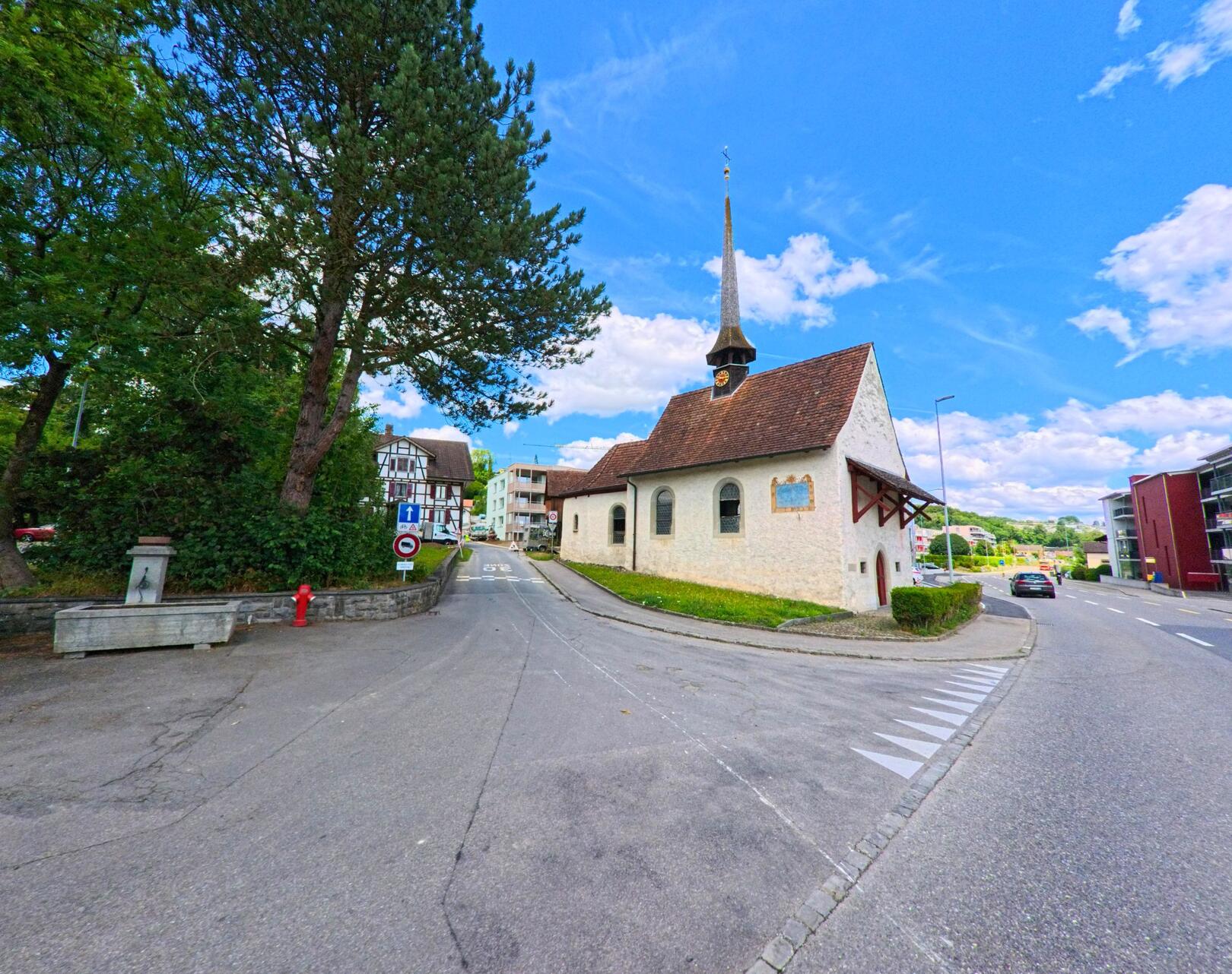 Die Kirche mit dem markanten Dachreiter steht im Zentrum von Geuensee (6232, Kanton Luzern) an einer kleinen Weggabelung, umgeben von traditionellen und modernen Gebaeuden.