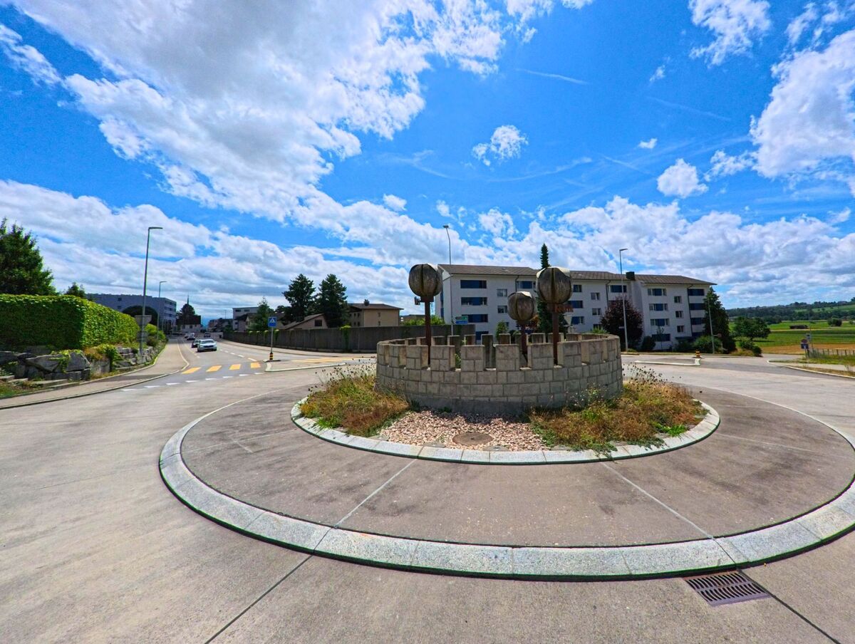 In einem Wohngebiet von Geuensee (6232, Kanton Luzern) liegt ein Kreisverkehr mit kuenstlerisch gestalteter Mittelinsel vor einem Mehrfamilienhaus und umgeben von Feldern.