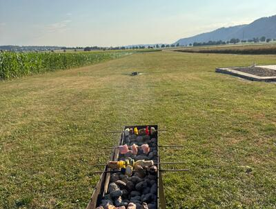 Auf einer Wiese in Fraubrunnen (3312, Kanton Bern) grillen Spiesschen ueber glühender Kohle, waehrend sich dahinter Felder und sanfte Huegel erstrecken.