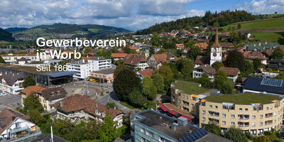 Über den Dächern von Worb [3076] im Kanton Bern zeigt das Bild eine weitläufige Ortsansicht mit Kirchturm, Wohnhäusern und bewaldeten Hügeln unter wolkigem Himmel.
