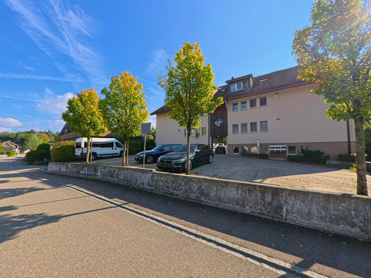 Vor einem grösseren Wohngebäude in Giebenach (4304, Kanton Basel-Landschaft) stehen mehrere Fahrzeuge neben einer Reihe junger Bäume entlang der Strasse.