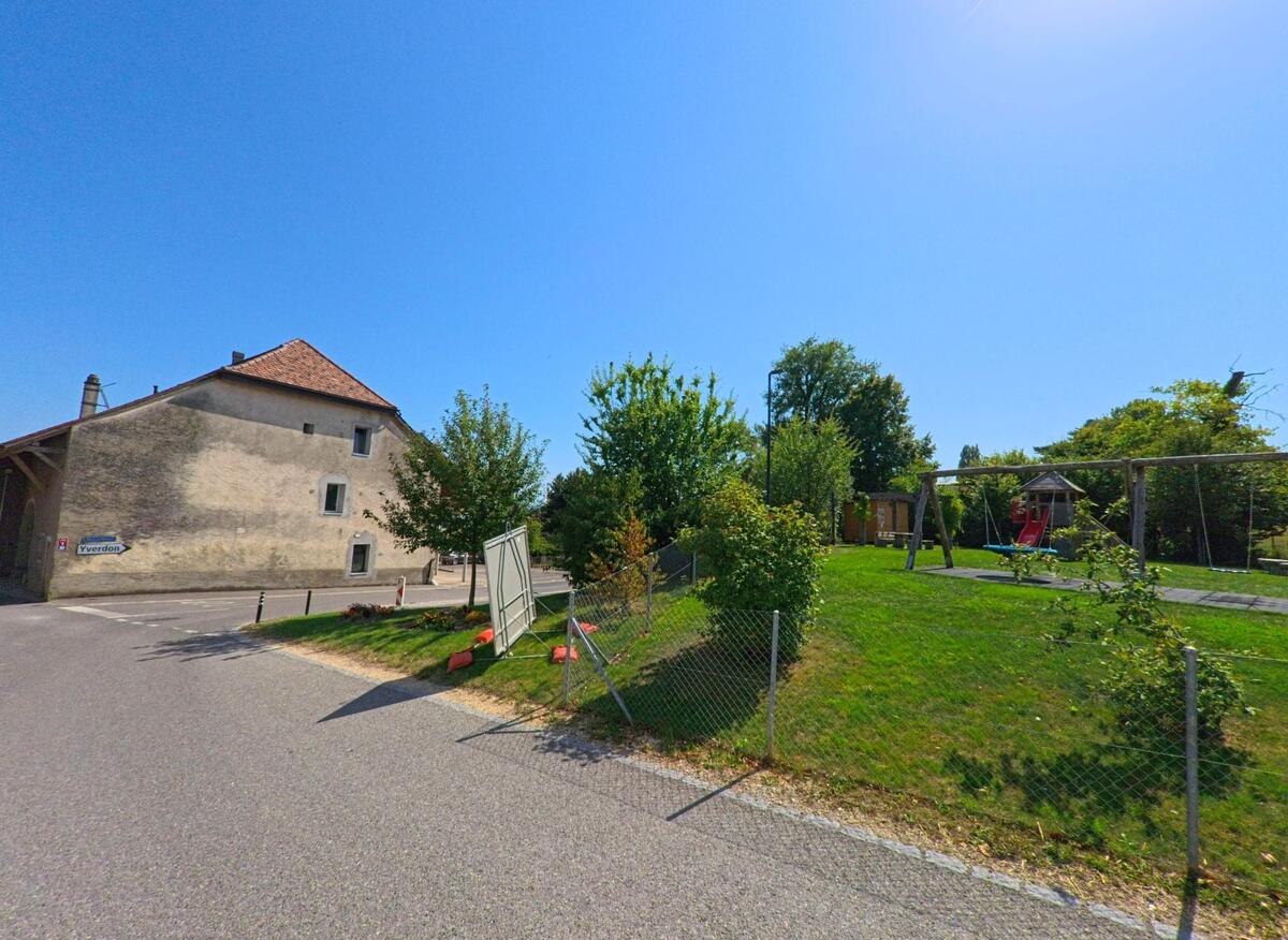 In Giez (1429, Kanton Waadt) steht ein altes Gebäude mit Ziegeldach neben einem eingezäunten Spielplatz mit Schaukeln und Bäumen.