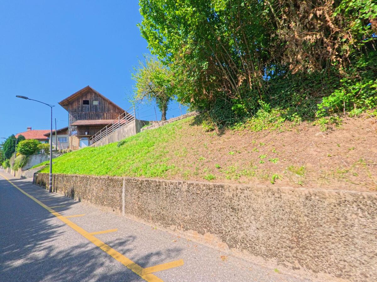 Ein traditionelles Holzhaus mit Balkon steht in Giez (1429, Kanton Waadt) oberhalb einer ansteigenden Strasse, die von einer Mauer und Grünstreifen begleitet wird.