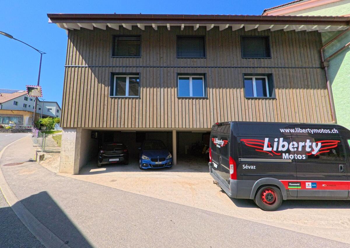 Ein modernes Wohnhaus mit Holzfassade steht in Giez (1429, Kanton Waadt), davor sind ein Lieferwagen und mehrere Autos unter einem Carport zu sehen.