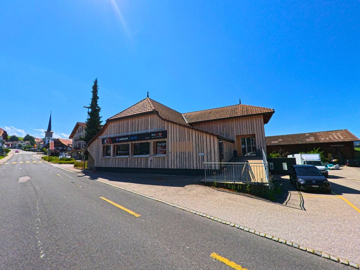 Entlang der Hauptstrasse in Giffers (1735, Kanton Freiburg) steht ein modernes Holzgebäude mit Geschäftsräumen, im Hintergrund ist die Kirche mit spitzem Turm sichtbar.
