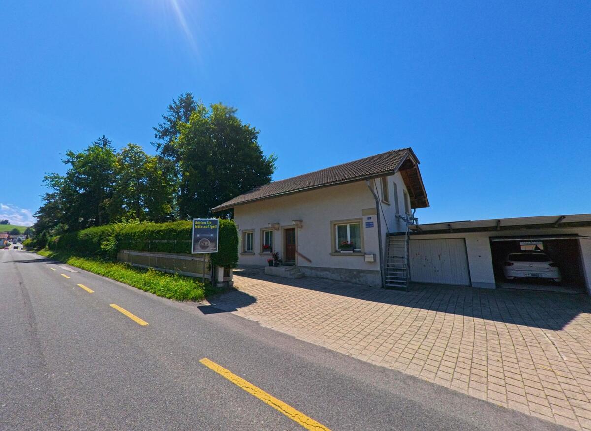 Ein Wohnhaus mit angebauter Garage steht an einer Strasse in Giffers (1735, Kanton Freiburg), vor einer Hecke mit einem Schild, das auf Igel hinweist.