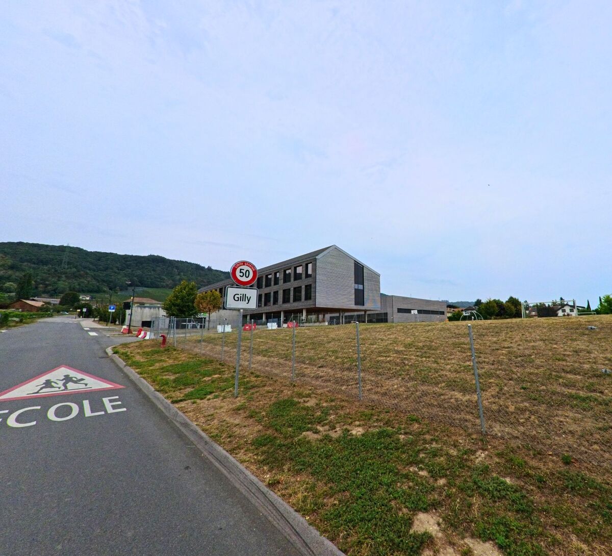 Am Ortseingang von Gilly (1182, Kanton Waadt) steht ein modernes Schulgebaeude neben dem Ortsschild und einem Tempolimit.