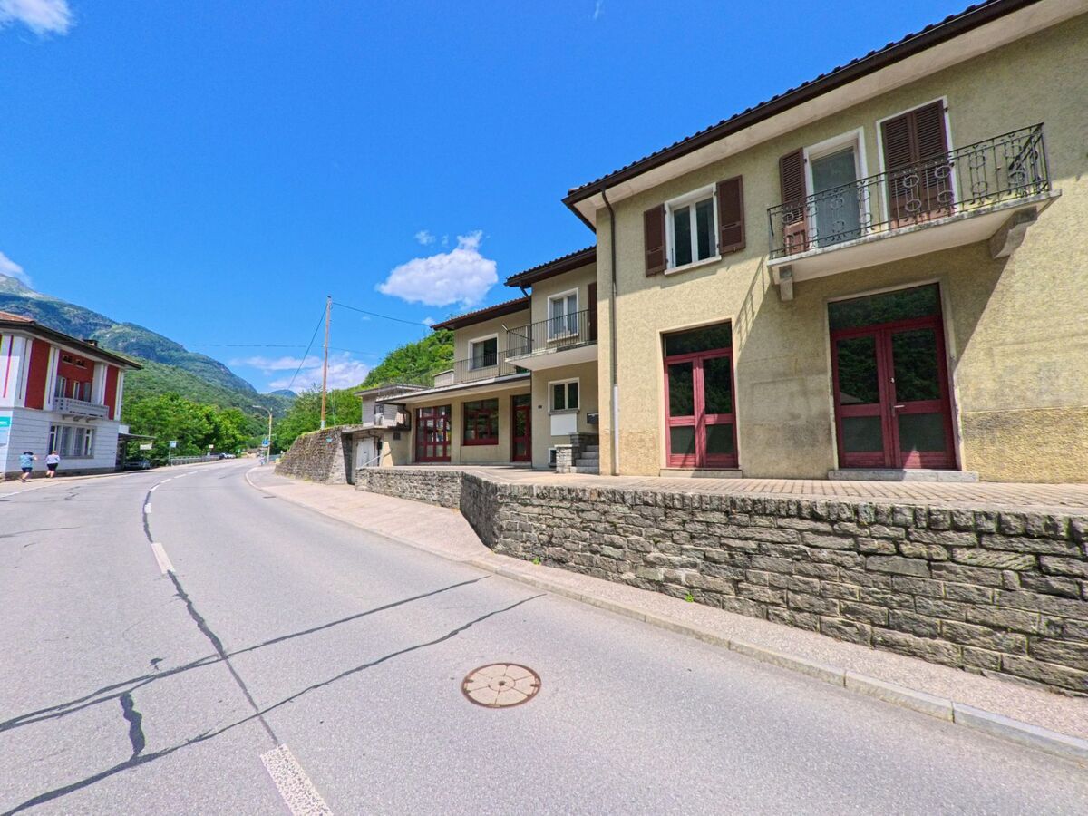 Entlang einer leicht gekruemmten Strasse in Giornico (6745, Kanton Tessin) stehen Haeuser mit Balkonen und roten Tueren vor einer Steinmauer, umgeben von gruenen Bergen.