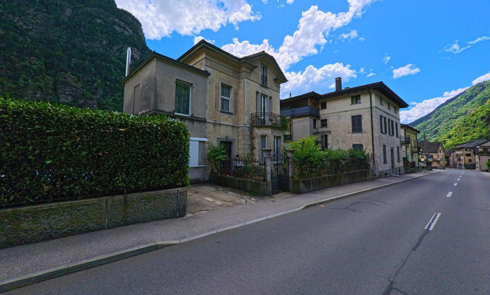 Entlang der Hauptstrasse in Giornico (6745, Kanton Tessin) stehen historische Wohnhaeuser mit gepflegten Gaerten und Blick auf die bewaldeten Berghaenge.