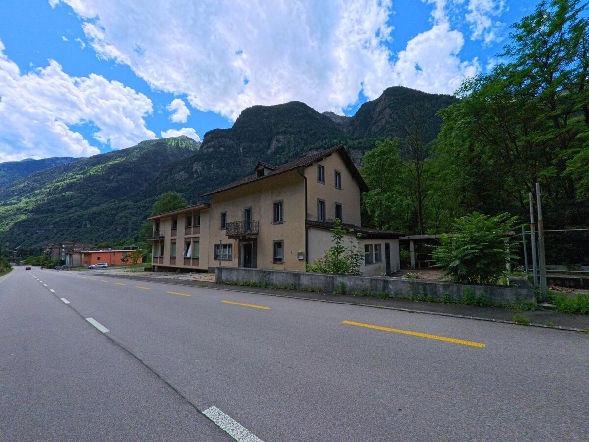 Ein zweistoeckiges Haus mit Balkon und umgebenden Baeumen steht in Giornico (6745, Kanton Tessin) an einer breiten Strasse mit Blick auf die gruenen Berghaenge.