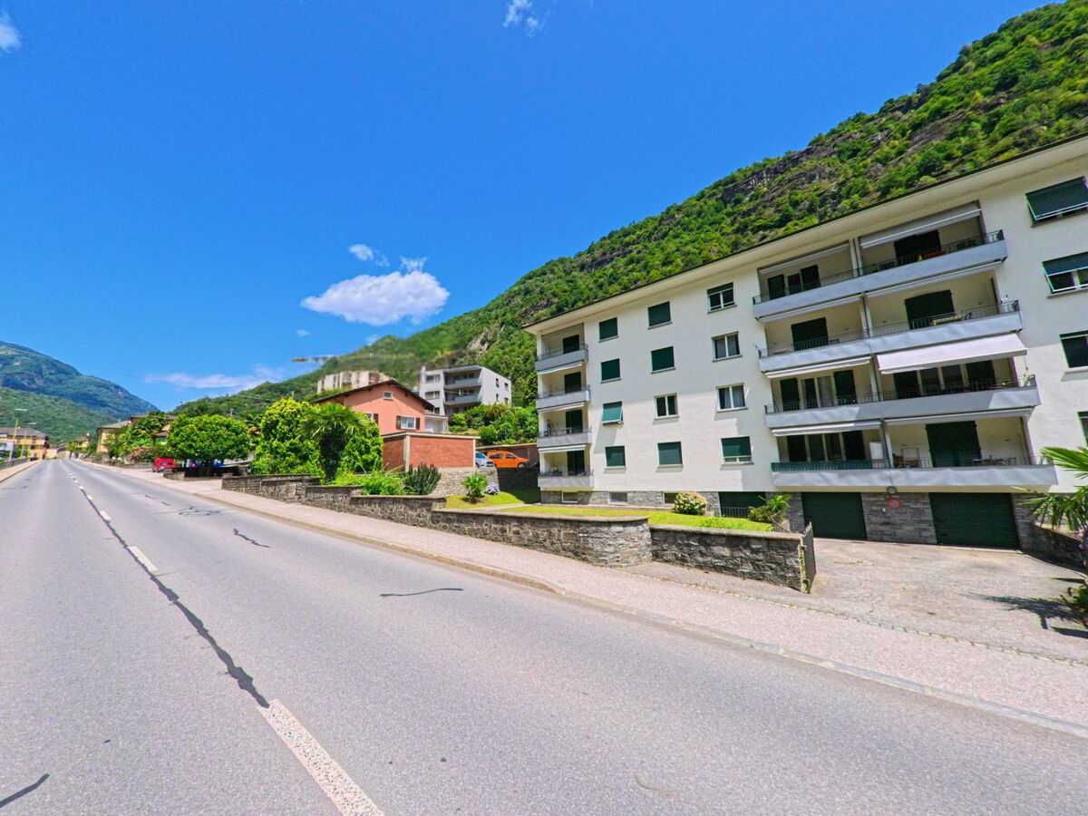 Mehrfamilienhaeuser mit Balkonen und gruene Gaerten saeumen in Giornico (6745, Kanton Tessin) die breite Hauptstrasse am Fusse der bewaldeten Haenge.