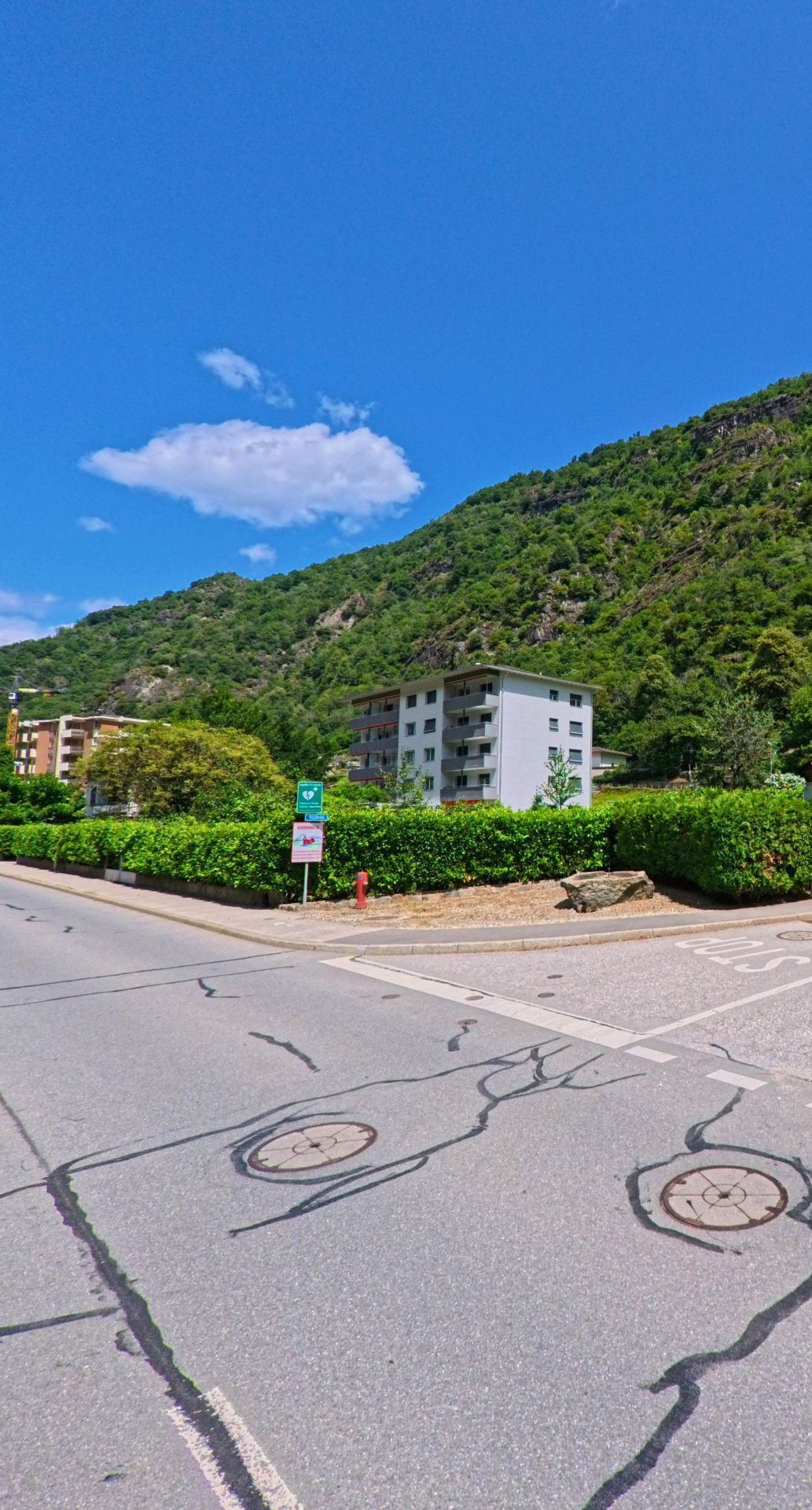 Ein modernes Mehrfamilienhaus in Giornico (6745, Kanton Tessin) liegt am Fuss eines bewaldeten Hangs, umgeben von gruener Hecke und Strassenmarkierungen im Vordergrund.