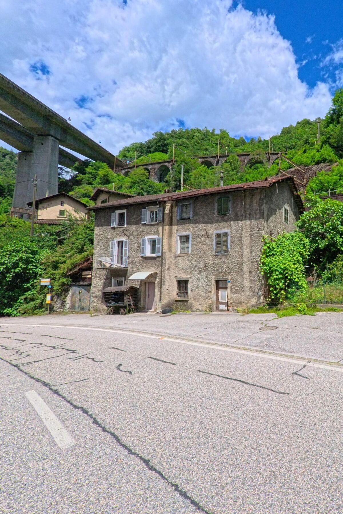Ein altes Steinhaus in Giornico (6745, Kanton Tessin) steht unterhalb einer imposanten Bruecke und eines Eisenbahnviadukts, umgeben von dichter Vegetation.