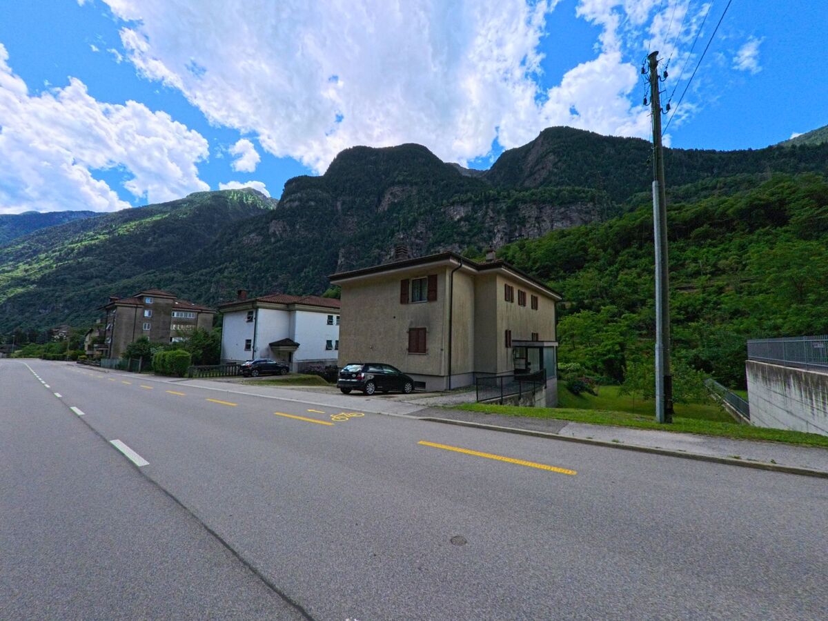 Am Rand von Giornico (6745, Kanton Tessin) verlaufen Wohnhaeuser entlang einer breiten Strasse, eingerahmt von den steilen, bewaldeten Haengen des Tals.