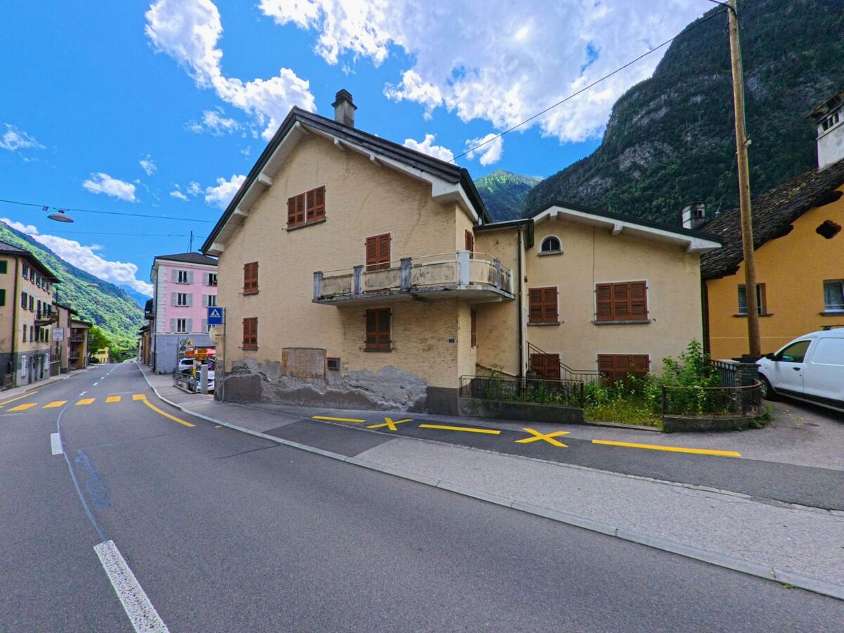 In Giornico (6745, Kanton Tessin) steht ein beiges Wohnhaus mit Holzladen und Balkon an einer Strassenkreuzung mit Blick auf die umliegenden Berge.