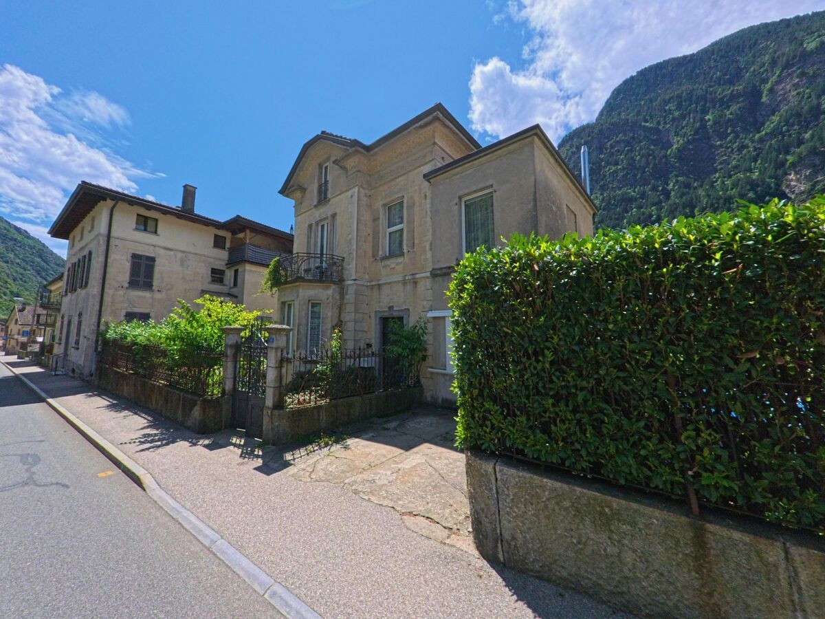 Ein helles Wohnhaus mit Balkon und gepflegtem Gartenzaun steht in Giornico (6745, Kanton Tessin) an einer schmalen Strasse vor den steilen Berghaengen.