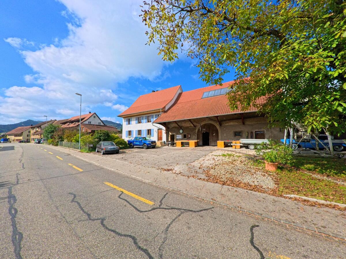 Ein traditionelles Bauernhaus mit rotem Dach und grossem Tor steht an einer sonnigen Strasse in Gipf-Oberfrick (5073, Kanton Aargau), daneben sind geparkte Fahrzeuge zu sehen.