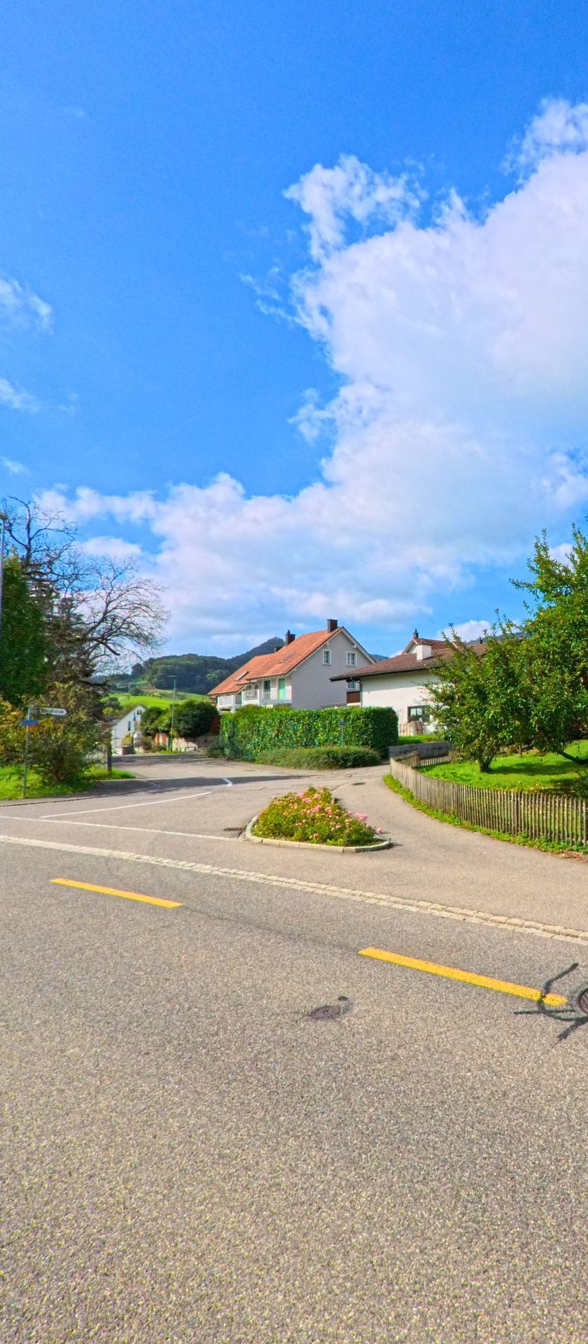 Eine ruhige Strasse in Gipf-Oberfrick (5073, Kanton Aargau) fuehrt an gepflegten Einfamilienhaeusern mit roten Daechern vorbei, umgeben von Gruenflaechen und huegeliger Landschaft.