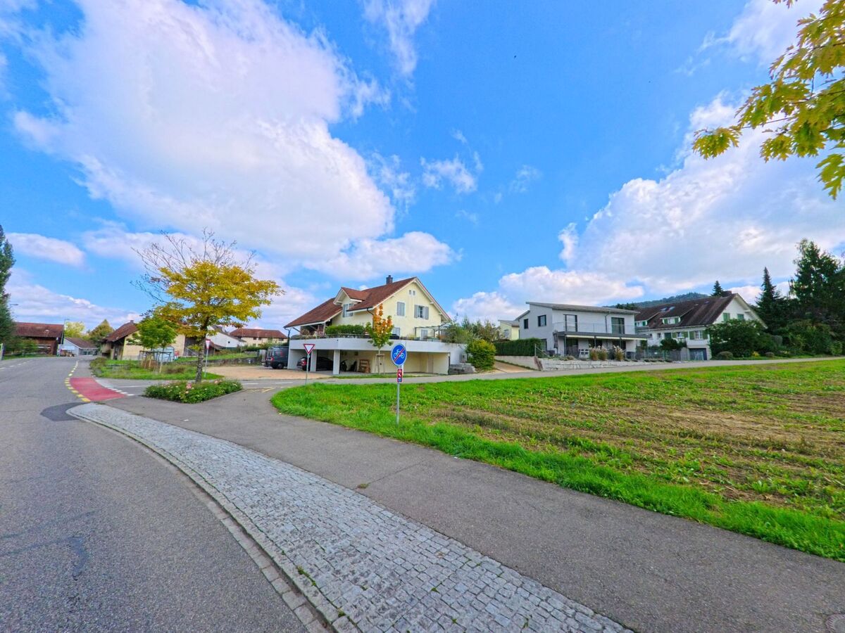 Am Ortsrand von Gipf-Oberfrick (5073, Kanton Aargau) stehen moderne und traditionelle Wohnhaeuser neben einem kleinen Feld unter blauem Himmel mit vereinzelten Wolken.