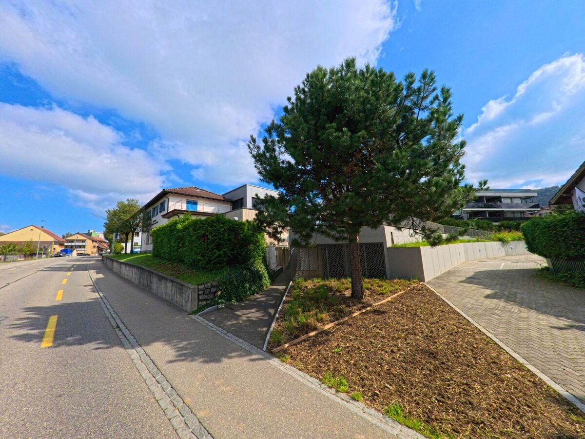 Eine leicht ansteigende Strasse in Gipf-Oberfrick (5073, Kanton Aargau) fuehrt an Wohnhaeusern mit Gaerten und einem einzelnen Baum im Vordergrund vorbei.