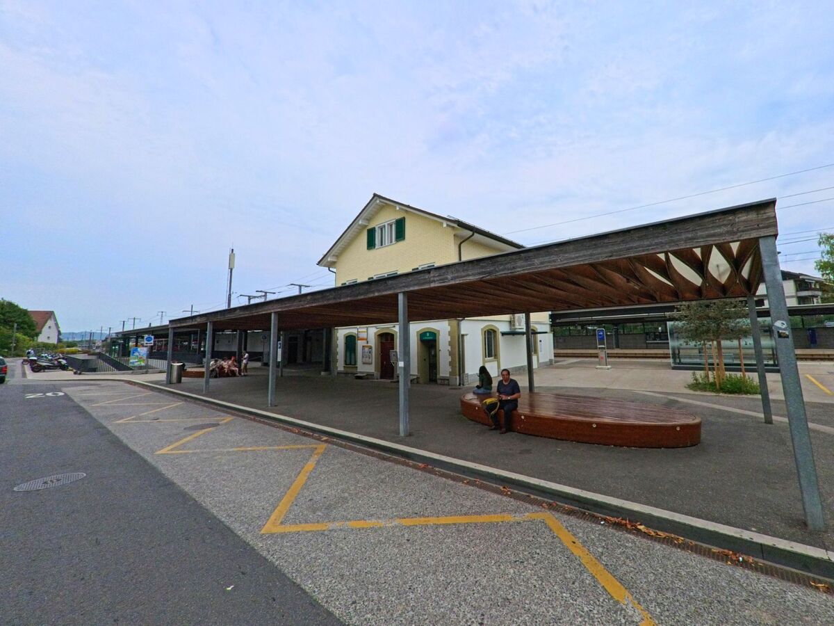 Der Bahnhof von Gland (1196, Kanton Waadt) zeigt ein gelbes Gebaeude mit Ueberdachung und Sitzgelegenheiten vor den Gleisen.