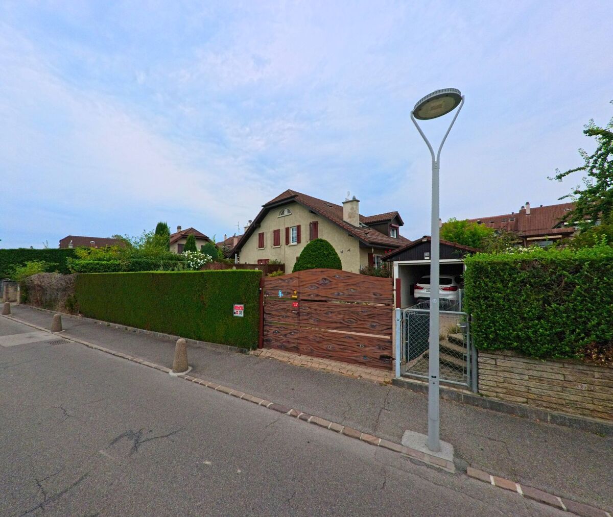 Ein Einfamilienhaus mit roten Fensterlaeden und grossem Holztor steht in einer Wohnstrasse von Gland (1196, Kanton Waadt).
