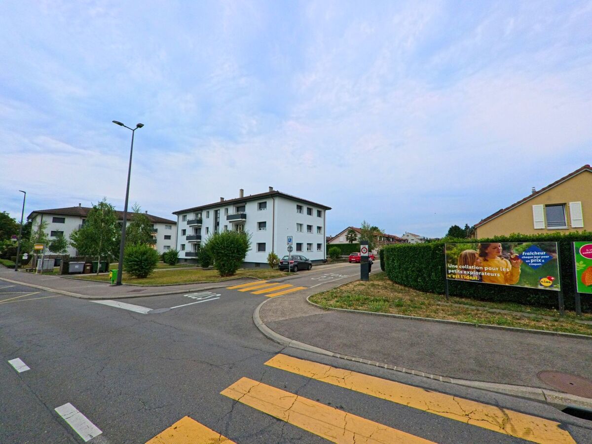 Mehrfamilienhaeuser mit Balkonen stehen an einer Strassenecke in Gland (1196, Kanton Waadt), neben einem Fussgaengerstreifen und Werbetafeln.