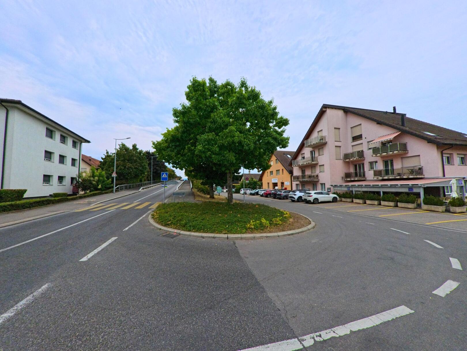 Eine Verkehrsinsel mit einem grossen Baum prägt den Strassenknotenpunkt in Gland (1196, Kanton Waadt), umgeben von Wohnhaeusern und einem Fussgaengerstreifen.
