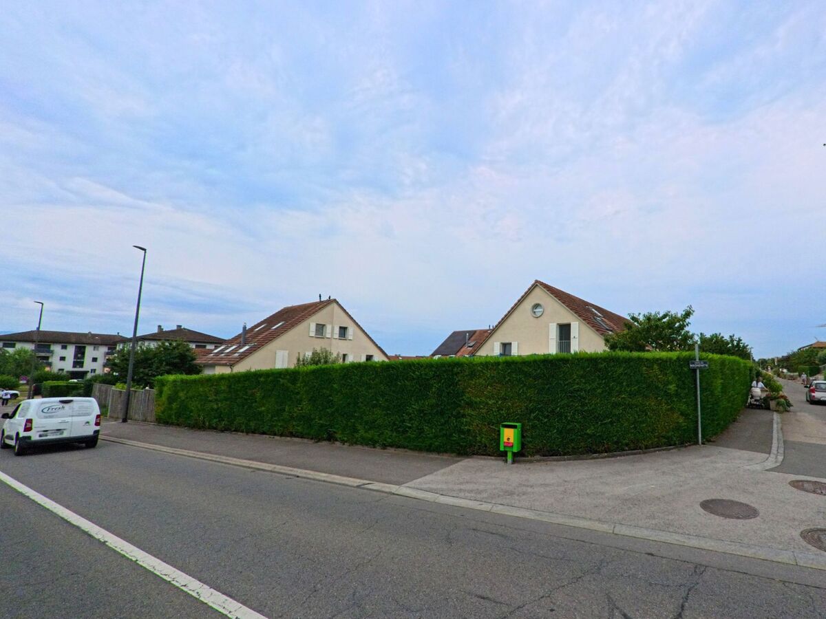 Zwei Wohnhaeuser mit roten Daechern liegen hinter einer dichten Hecke an einer Strasse in Gland (1196, Kanton Waadt).