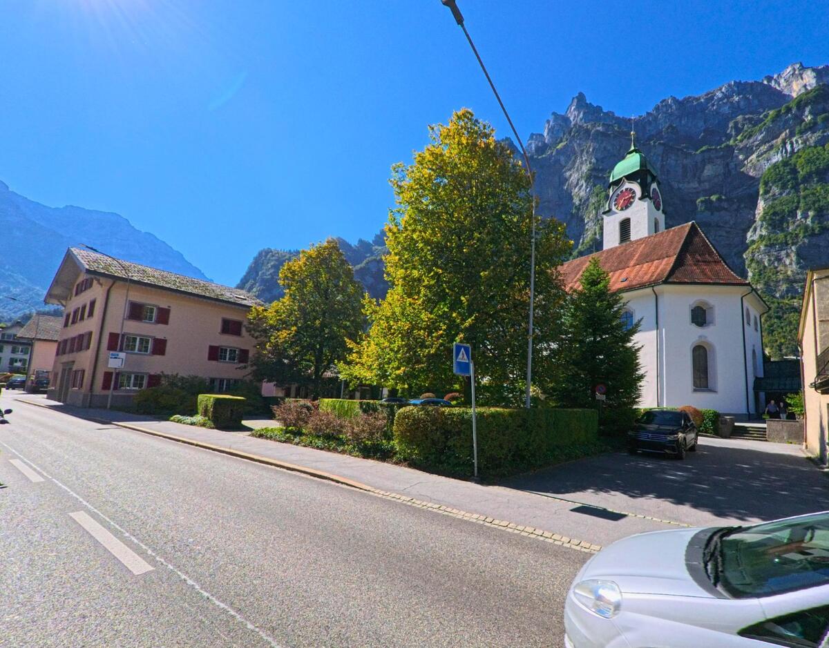 Die reformierte Kirche in Glarus (8754, Kanton Glarus) steht an einer sonnigen Strasse, eingerahmt von Bergen und grünen Bäumen.