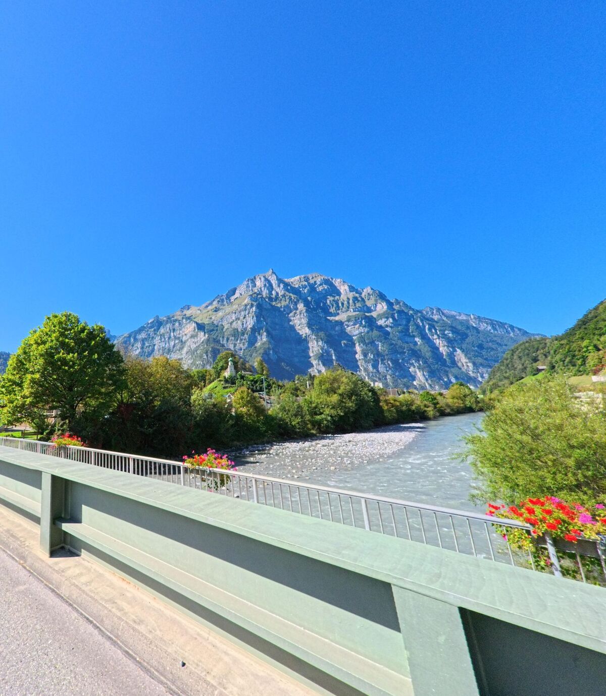 Von einer Bruecke in Glarus (8755, Kanton Glarus) bietet sich ein klarer Blick auf den Fluss und den markanten Berg im Hintergrund unter blauem Himmel.
