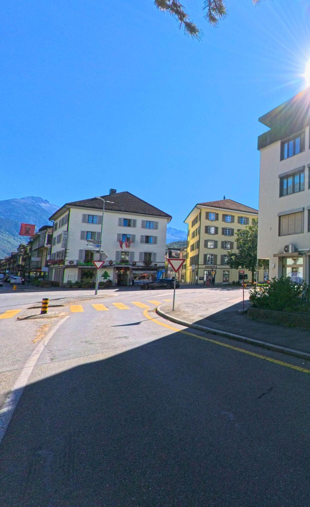 Eine sonnige Strassenszene in Glarus (8755, Kanton Glarus) zeigt eine Kreuzung mit Fussgaengerstreifen und mehreren Wohnhaeusern vor alpiner Kulisse.