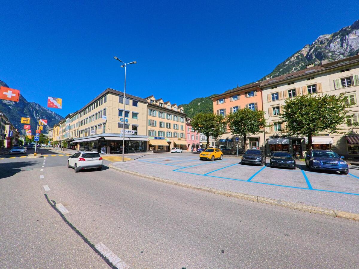 In der Sonne zeigt sich das Zentrum von Glarus (8755, Kanton Glarus) mit seinen bunt gestrichenen Fassaden, geparkten Autos und den umliegenden Bergen im Hintergrund.