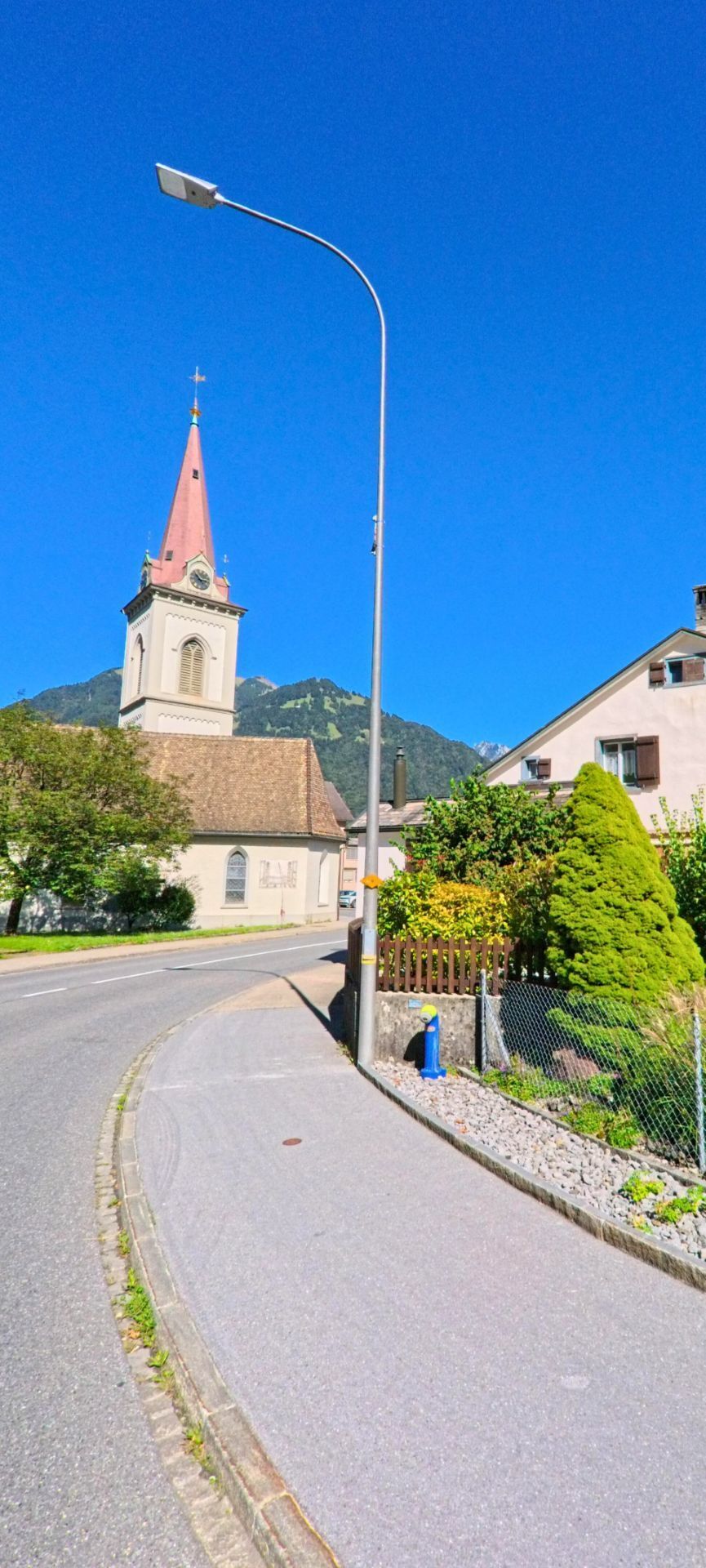 Entlang eines schmalen Gehwegs führt die Strasse an einer Kirche mit Turm vorbei, die das Ortsbild von Glarus Nord (8865, Kanton Glarus) vor der Bergkulisse prägt.