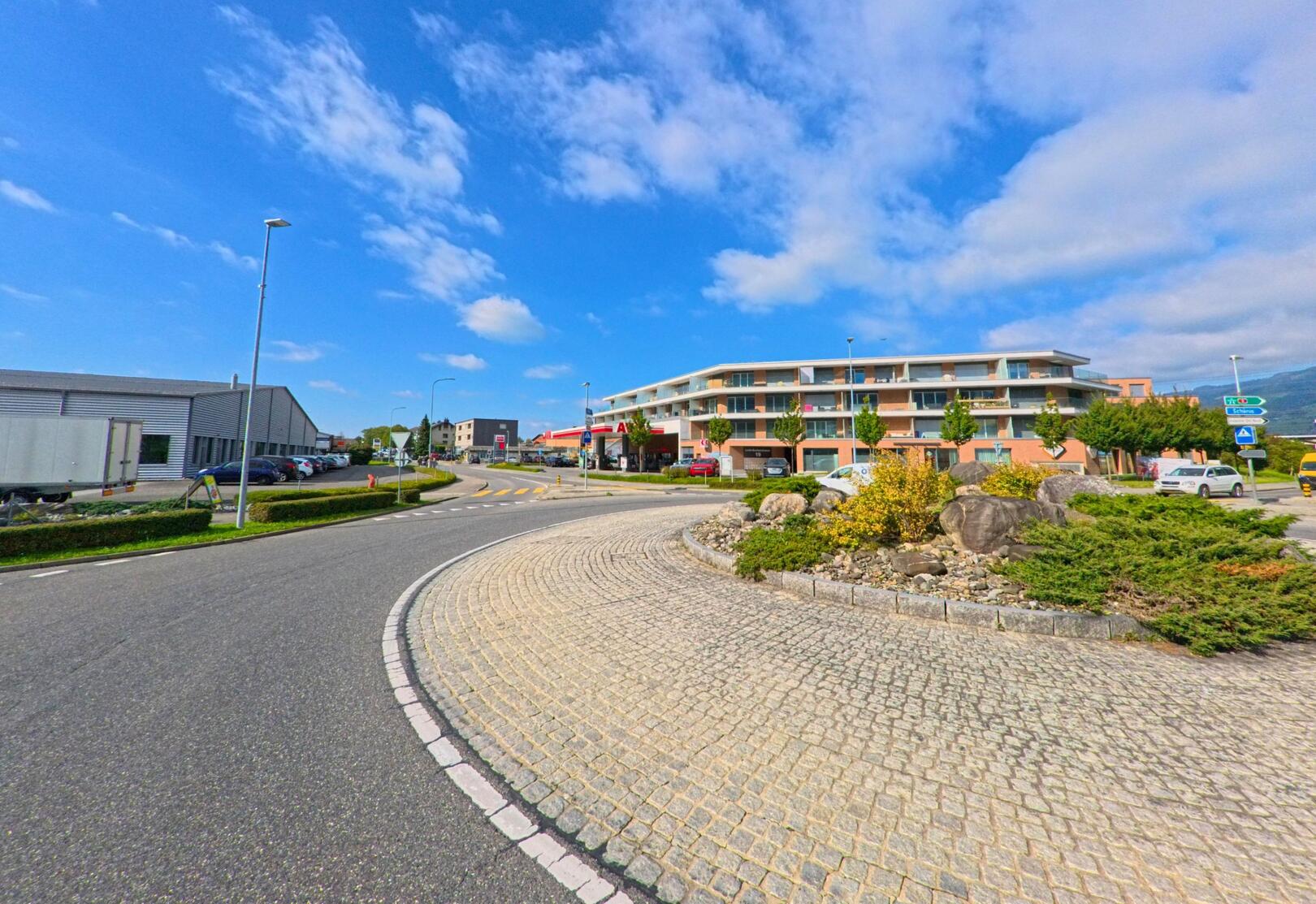 An einer gepflegten Verkehrsinsel mit Natursteinen und Bepflanzung verläuft ein Kreisverkehr in Glarus Nord (8865, Kanton Glarus), umgeben von modernen Wohn- und Gewerbebauten unter blauem Himmel.