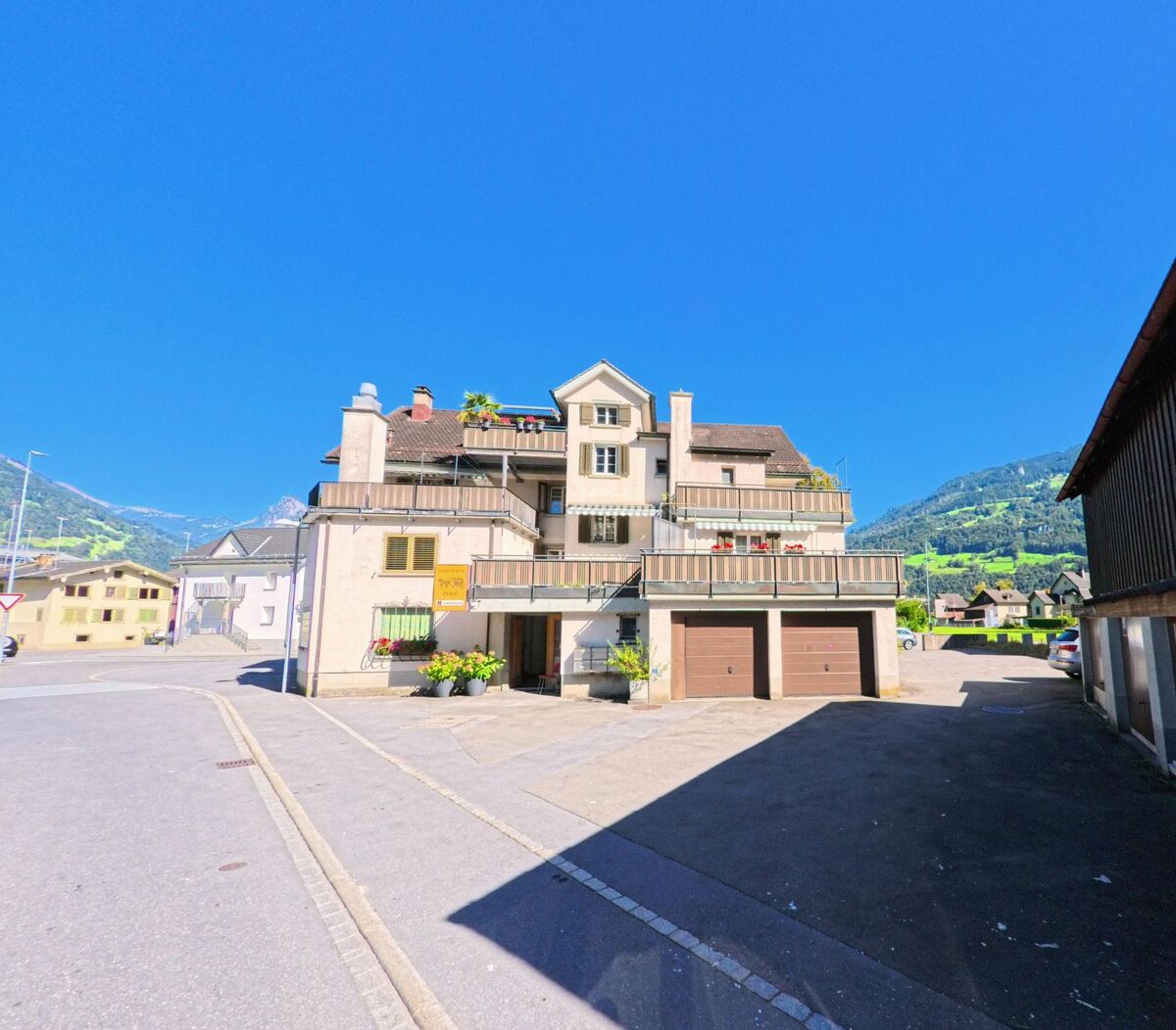 An einem offenen Platz steht ein mehrgeschossiges Wohnhaus mit Balkonen und Garagentoren in Glarus Nord (8865, Kanton Glarus), eingerahmt von Nachbarbauten und der umliegenden Berglandschaft.